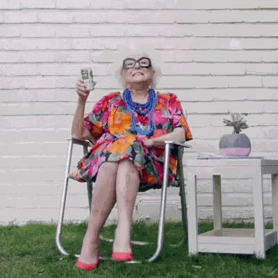 Stará žena v barevných šatech, sedící na židli a připíjející na zdraví.