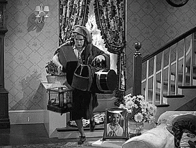 #PRATODOSVEREM: Uma cena em preto e branco de uma mulher segurando malas e bolsas nas duas mãos e correndo com dificuldade na sala, em direção à porta.