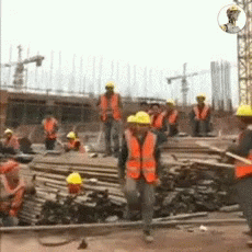 A person in a bright orange safety vest and hard hat dancing in front of a crew of construction workers at a job site with cranes visible in the background.